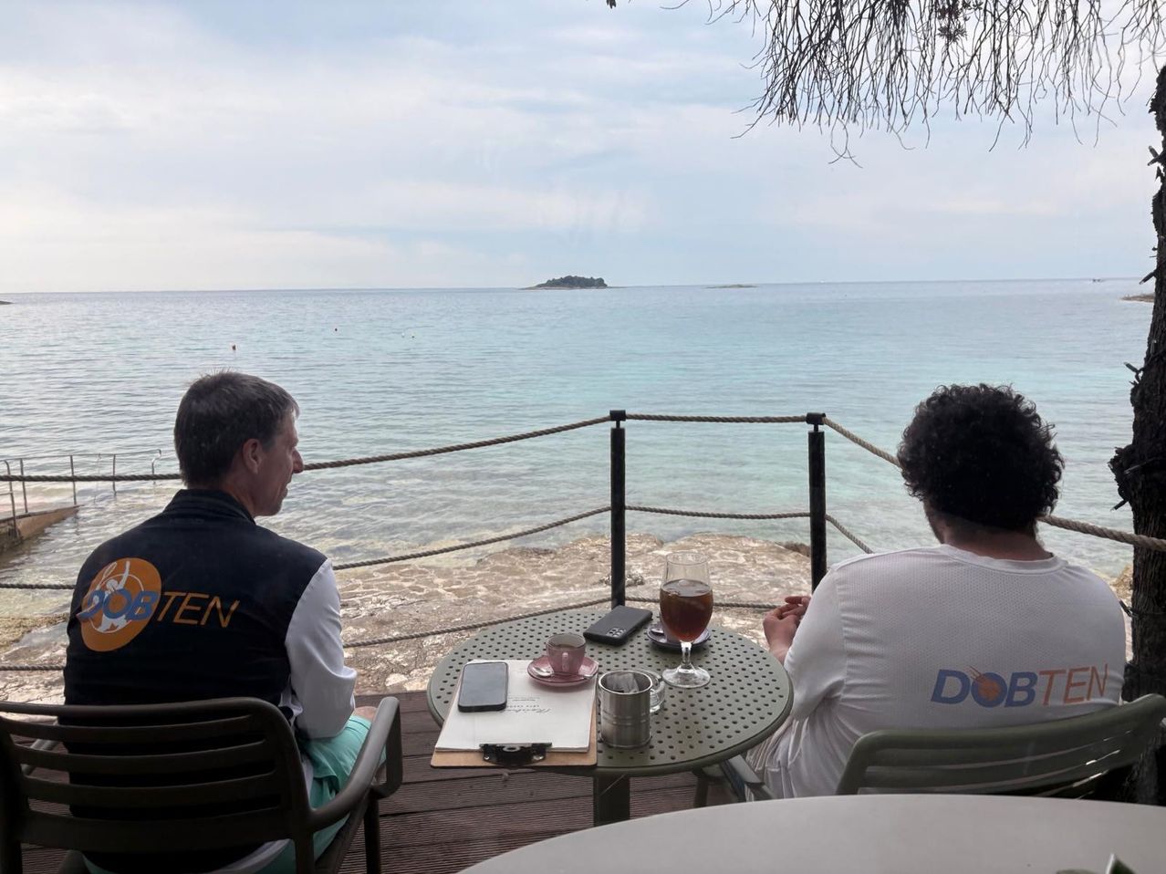 Zwei Personen sitzen auf einem Balkon mit Meerblick, eine trägt eine Weste und die andere ein weißes T-Shirt. Ein Tisch zwischen ihnen hat ein Glas, eine Tasse, ein Handy und ein Notizbuch. In der Ferne ist eine kleine Insel zu sehen.