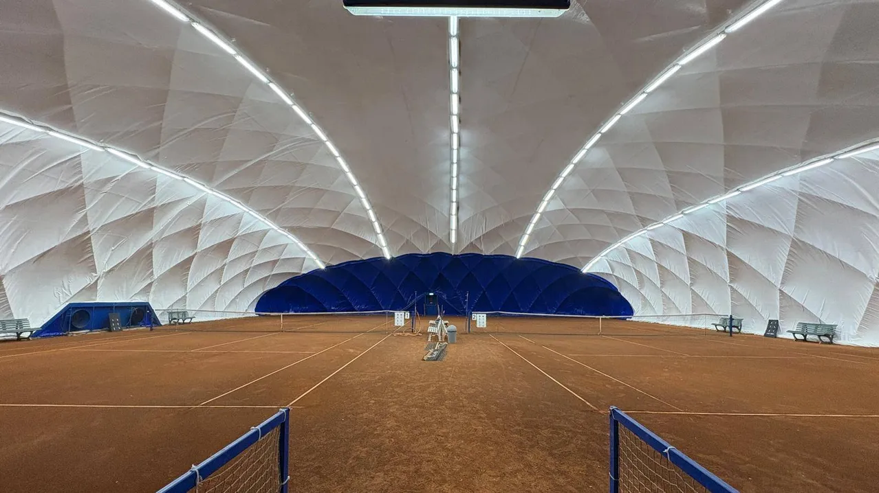 Ein Indoor-Tennisplatz mit einer weißen und blauen Zeltstruktur, mit einer blauen Wand im Hintergrund und weißen Linien auf dem Boden.