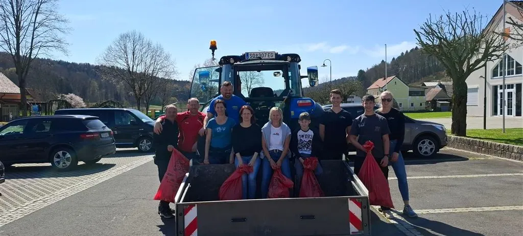 Eine Gruppe von Menschen posiert für ein Foto neben einem blauen Traktor mit roten Beuteln im Heck. Die Umgebung ist ein offener Parkplatz mit geparkten Autos und Bäumen im Hintergrund.