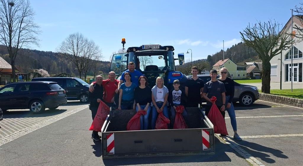 Eine Gruppe von Menschen posiert für ein Foto neben einem blauen Traktor mit roten Beuteln im Heck. Die Umgebung ist ein offener Parkplatz mit geparkten Autos und Bäumen im Hintergrund.