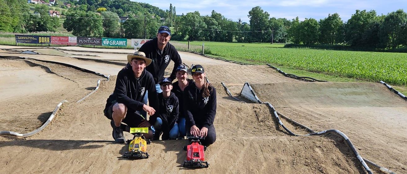 Eine Familie posiert auf einer Sandrennstrecke mit Bäumen und einem Feld im Hintergrund. Sie halten Spielzeugautos in den Händen.