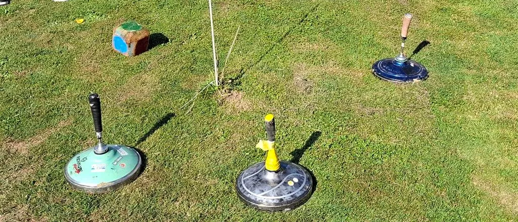 Ein Außenbereich mit Rasenfläche hat vier Rasenmäher, von denen einer gelb und schwarz bemalt ist. Ein kleiner Holzkasten ist ebenfalls vorhanden.