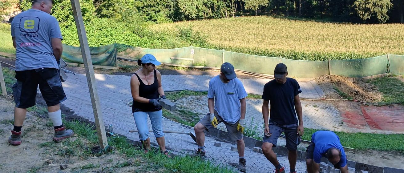 Eine Gruppe von Menschen arbeitet an einem Pfad in einem Grasfeld, einer benutzt einen Spaten und ein anderer hält einen Ziegelstein. Bäume und ein Zaun sind im Hintergrund.