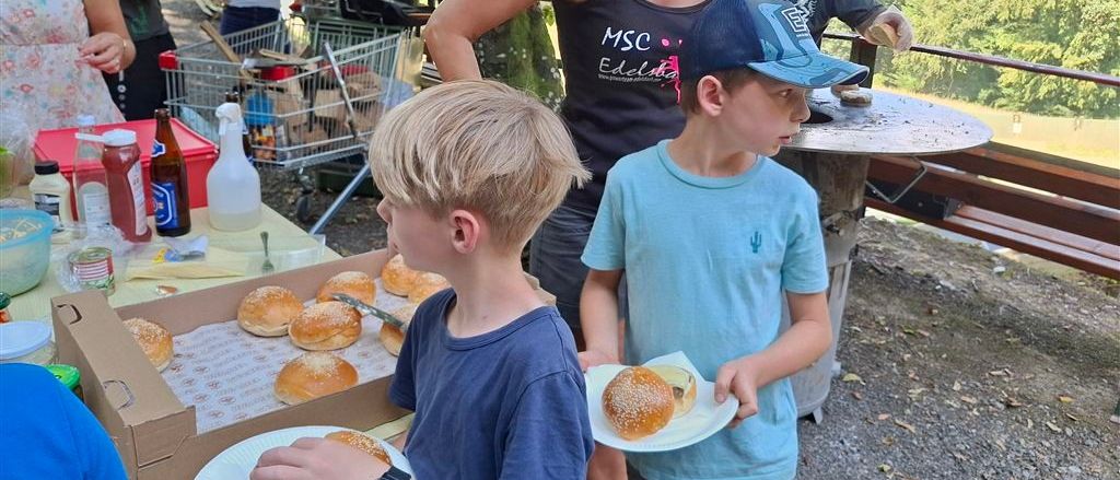Eine Familie versammelt sich draußen mit einem Tisch voller Sandwiches und Getränken. Eine Frau hält einen Hamburger und ein Junge ein Sandwich. Ein weiterer Junge und ein Mann sind im Hintergrund.