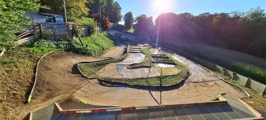 Eine Luftaufnahme einer Kurvenstrecke aus Erde mit Rampen und Pfützen, vor einem Hintergrund aus üppigem Grün und einem strahlenden Sonnenschein. Unten befindet sich eine Holzbarriere mit roten und weißen Streifen.