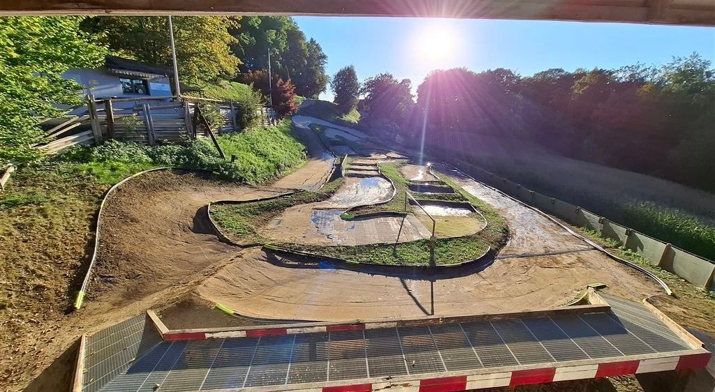 Eine Luftaufnahme einer Kurvenstrecke aus Erde mit Rampen und Pfützen, vor einem Hintergrund aus üppigem Grün und einem strahlenden Sonnenschein. Unten befindet sich eine Holzbarriere mit roten und weißen Streifen.