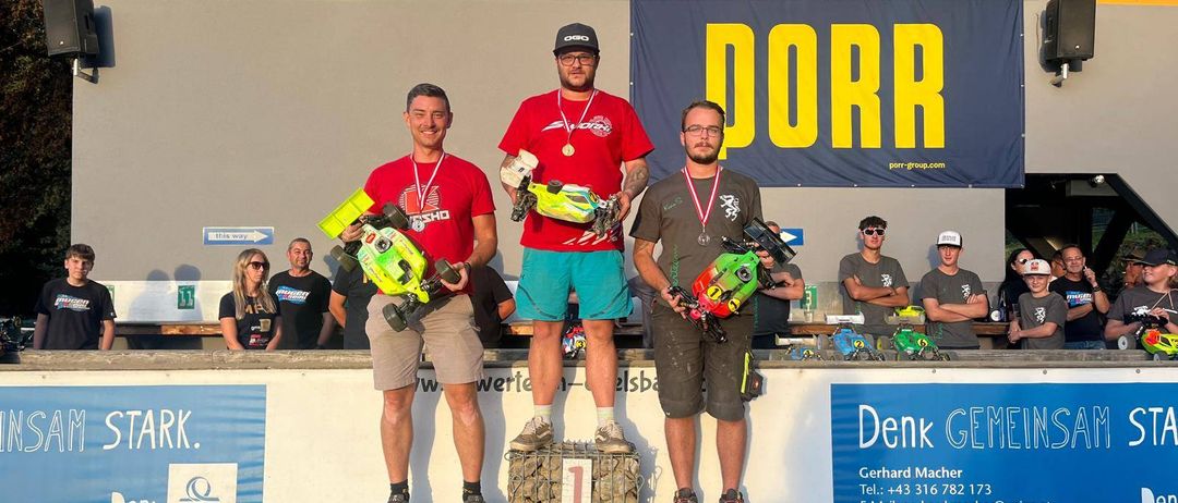 Drei Männer stehen auf einem Podest mit ferngesteuerten Autos und Medaillen. Das Podest hat die Zahlen 1, 2 und 3. Sie befinden sich bei einer Outdoor-Veranstaltung mit Bannern dahinter.