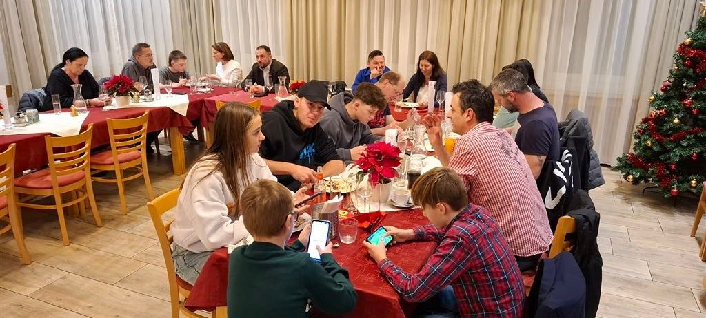 Eine Gruppe von Menschen sitzt um einen Esstisch herum, einige führen Gespräche, andere nutzen ihre Handys. Der Tisch ist mit Tellern, Tassen und Blumen gedeckt. Im Hintergrund hängen weiße Vorhänge.