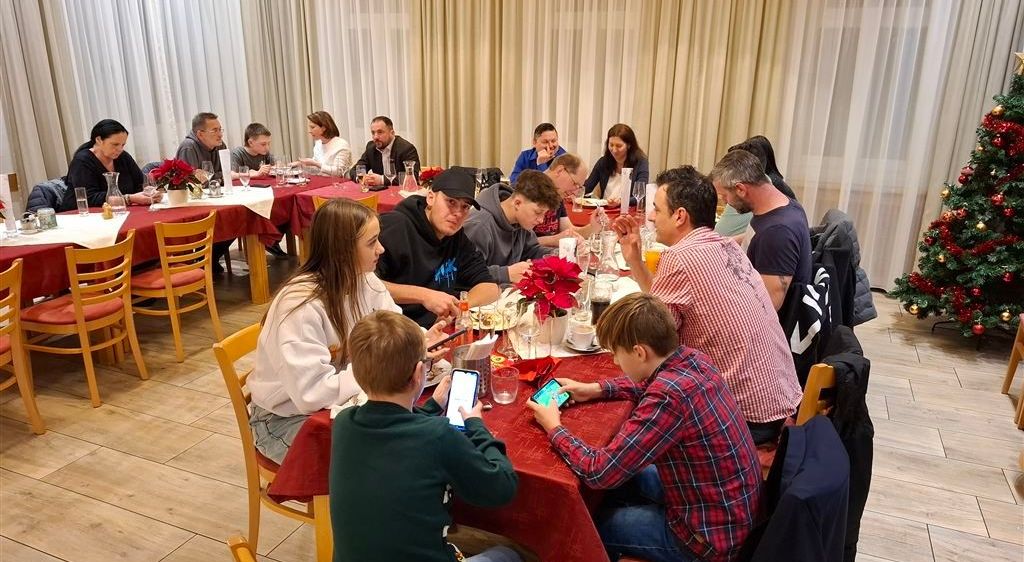 Eine Gruppe von Menschen sitzt um einen Esstisch herum, einige führen Gespräche, andere nutzen ihre Handys. Der Tisch ist mit Tellern, Tassen und Blumen gedeckt. Im Hintergrund hängen weiße Vorhänge.