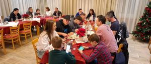 Eine Gruppe von Menschen sitzt um einen Esstisch herum, einige führen Gespräche, andere nutzen ihre Handys. Der Tisch ist mit Tellern, Tassen und Blumen gedeckt. Im Hintergrund hängen weiße Vorhänge.