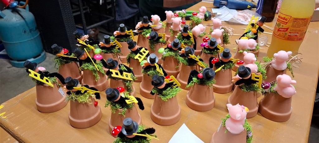 Ein Holztisch hat mehrere Tontöpfe mit grünem Moos und kleinen Figuren darauf, einige tragen Hüte und halten Hämmer.