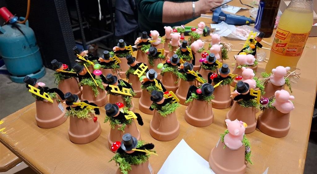 Ein Holztisch hat mehrere Tontöpfe mit grünem Moos und kleinen Figuren darauf, einige tragen Hüte und halten Hämmer.