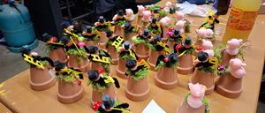 Ein Holztisch hat mehrere Tontöpfe mit grünem Moos und kleinen Figuren darauf, einige tragen Hüte und halten Hämmer.