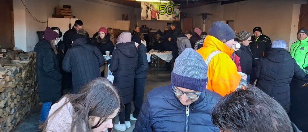 Eine Gruppe von Menschen in Winterkleidung steht in einem Raum, wahrscheinlich zur Feier eines Geburtstags. Ein Banner mit der Zahl '50' hängt an der Wand.