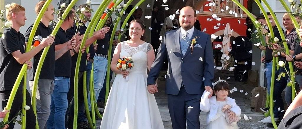 Eine Hochzeitsfeier unter einem Bogen aus grünen Röhren mit fallenden Blütenblättern, die Braut und der Bräutigam halten sich an den Händen, und ein kleines Mädchen geht neben ihnen.