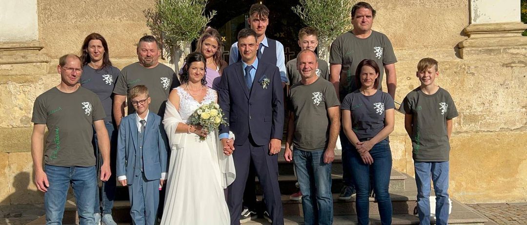 Eine Hochzeitsgesellschaft posiert auf einer Treppe mit steinernem Hintergrund, alle in aufeinander abgestimmten Outfits, mit der Braut, dem Bräutigam und mehreren Familienmitgliedern.