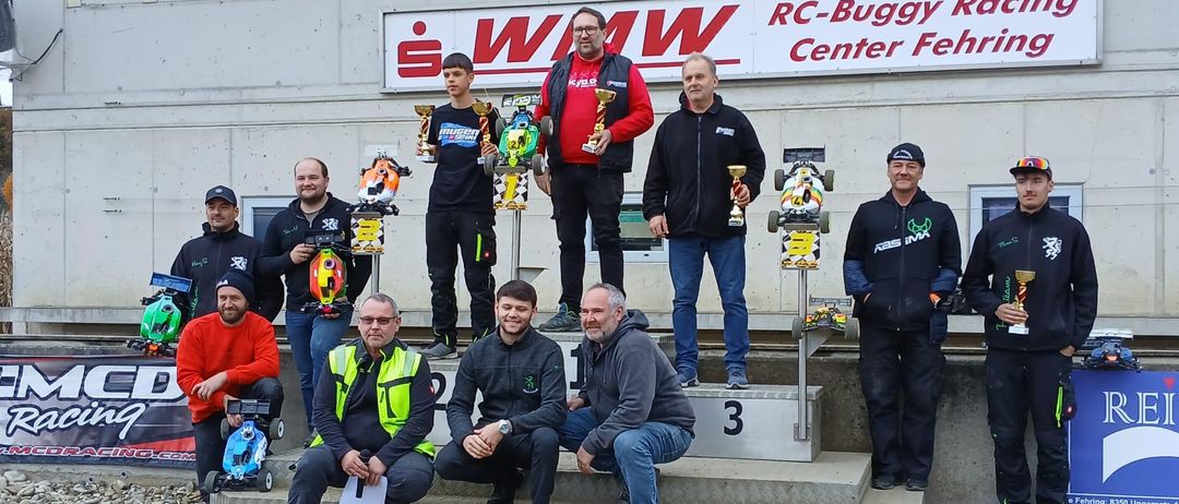 Mehrere Männer stehen auf einem Podest und halten Trophäen und ferngesteuerte Autos. Hinter ihnen steht ein Gebäude mit einem Schild, das 'RC-Buggy Racing Center Fehringen' besagt.