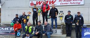 Mehrere Männer stehen auf einem Podest und halten Trophäen und ferngesteuerte Autos. Hinter ihnen steht ein Gebäude mit einem Schild, das 'RC-Buggy Racing Center Fehringen' besagt.
