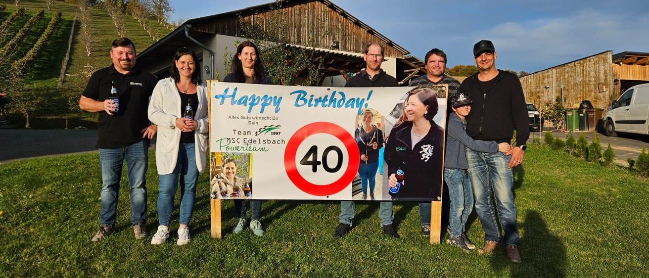 Eine Gruppe von Menschen feiert einen Geburtstag mit einem Banner, das 'Happy Birthday!' sagt, mit einem Nummer-40-Schild und Bildern von Menschen. Sie stehen vor einem Haus mit einem Bauernhaus.
