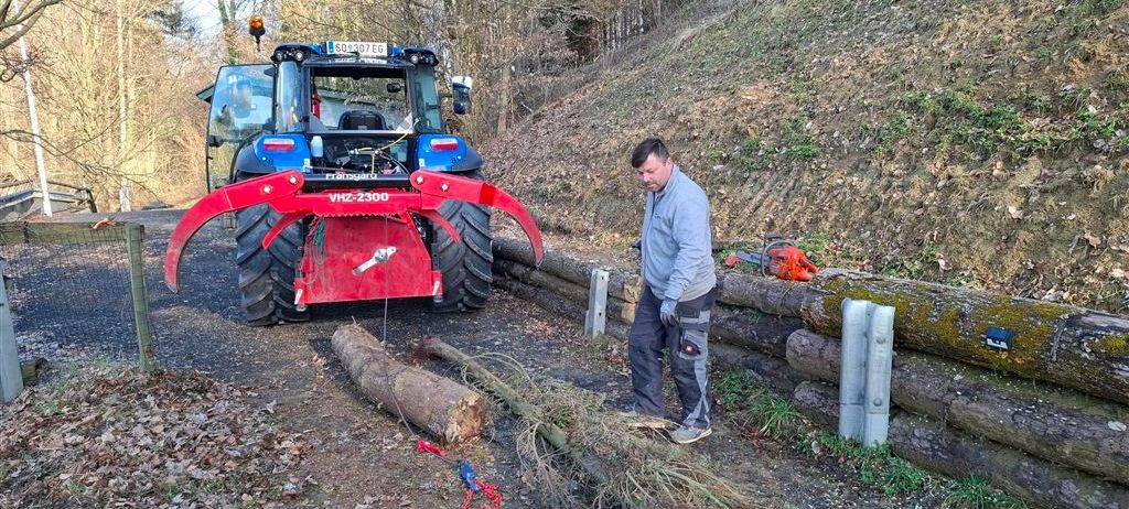Ein Mann in einem grauen Jacken arbeitet im Freien neben einem blauen Traktor mit einem roten Anbau. Er steht neben einem Baumstamm und Zweigen mit Ketten.