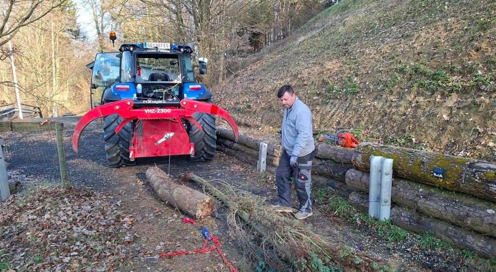 Ein Mann in einem grauen Jacken arbeitet im Freien neben einem blauen Traktor mit einem roten Anbau. Er steht neben einem Baumstamm und Zweigen mit Ketten.