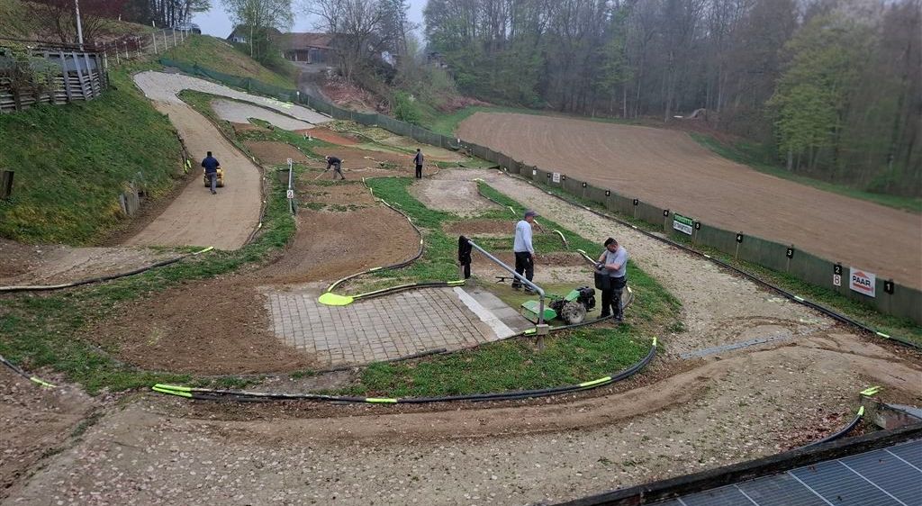 Eine Dirtbike-Strecke mit arbeitenden Personen, einer, der ein Fahrrad fährt, und anderen, die stehen und Werkzeuge halten.