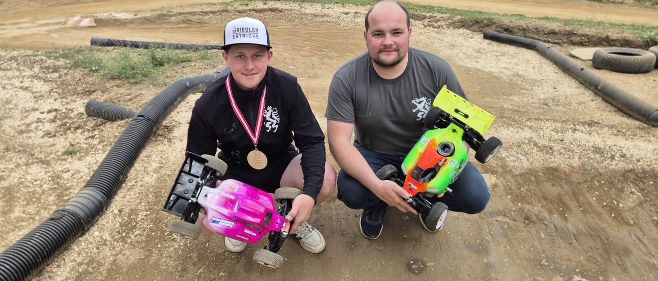 Zwei Männer posieren mit ihren Spielzeugautos auf einer Erdstrecke, einer trägt eine Medaille. Hinter ihnen befinden sich Zäune mit Bannern und einem Hintergrund aus Hügeln.