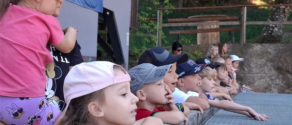 Eine Gruppe von Kindern mit Mützen schaut sich eine Veranstaltung im Freien an, einige mit den Händen am Mund. Eine Frau mit Sonnenbrille steht hinter ihnen.