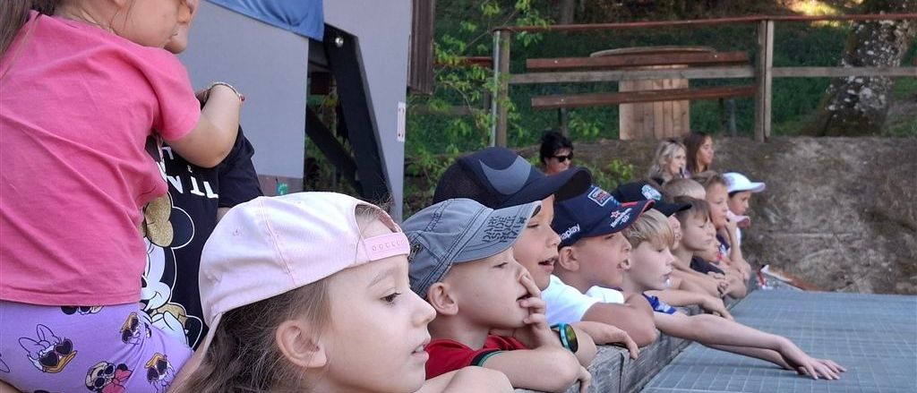 Eine Gruppe von Kindern mit Mützen schaut sich eine Veranstaltung im Freien an, einige mit den Händen am Mund. Eine Frau mit Sonnenbrille steht hinter ihnen.