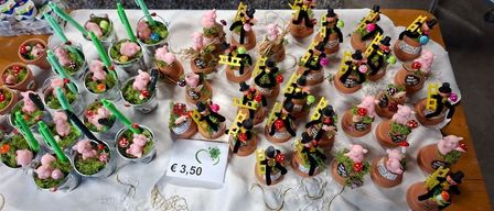 Ein Tisch zeigt mehrere Terrakotta-Töpfe mit Schweinfiguren, jeweils begleitet von grünen Strohhalmen und einem Preisschild von 3,50 €. Im Hintergrund befinden sich ein grauer Teppich und ein silbernes Metallobjekt.