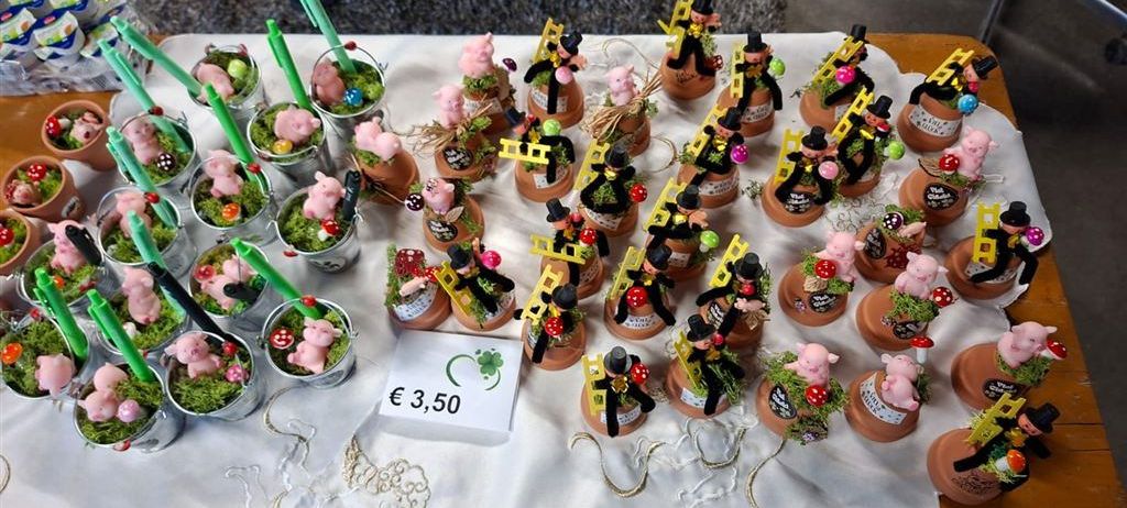 Ein Tisch zeigt mehrere Terrakotta-Töpfe mit Schweinfiguren, jeweils begleitet von grünen Strohhalmen und einem Preisschild von 3,50 €. Im Hintergrund befinden sich ein grauer Teppich und ein silbernes Metallobjekt.