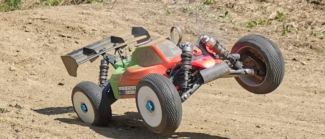 Ein ferngesteuertes Offroad-Rennauto mit orange-grünem Körper, weißen Rädern und schwarzen Reifen, auf einer Erderpiste geparkt.