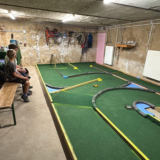 Drei Kinder sitzen auf einer Bank und beobachten eine Rennstrecke mit einem Spielzeugauto. Die Strecke ist grün mit gelben Markierungen.