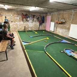 Drei Kinder sitzen auf einer Bank und beobachten eine Rennstrecke mit einem Spielzeugauto. Die Strecke ist grün mit gelben Markierungen.