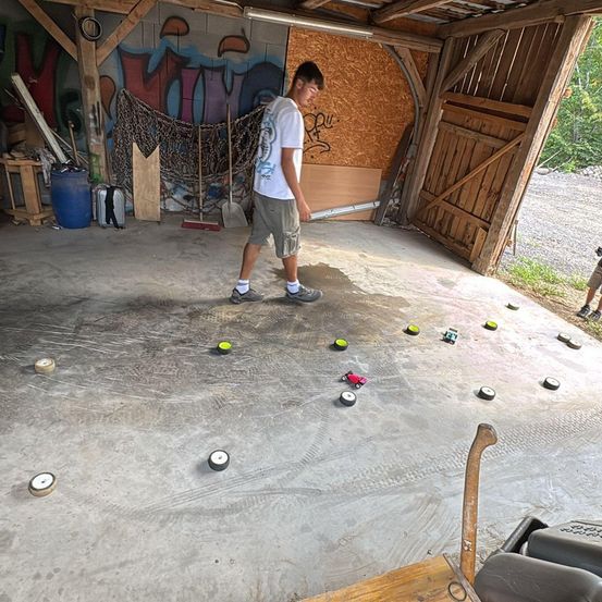 Ein Mann geht in einer Garage mit Graffiti an der Wand, hält einen Stock und ist von kleinen Objekten umgeben. Er trägt Turnschuhe. Ein blauer Eimer steht auf dem Boden.