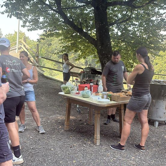 Eine Gruppe von Menschen steht unter einem Baum und bereitet Essen auf einem Tisch zu. Ein Mann hält ein Bier, während andere sich um ihn versammeln. Ein Trolley mit Essen in der Nähe.