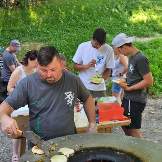 Ein Mann kocht Burger auf einem Grill im Freien. Mehrere Menschen versammeln sich, essen und unterhalten sich. Ein Tisch hält Essen, Getränke und eine Aufbewahrungsbox.