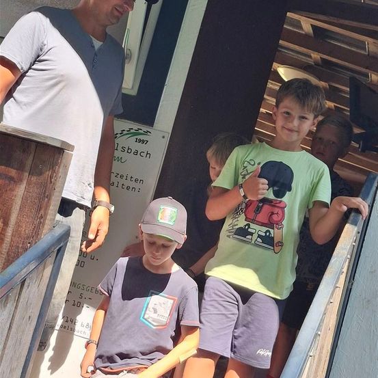 Eine Gruppe von Jungen und einem Mann sind auf einer Treppe, wobei ein Junge den Daumen hochhält. Sie verlassen möglicherweise ein Gebäude mit einem Schild mit der Aufschrift '1997'.