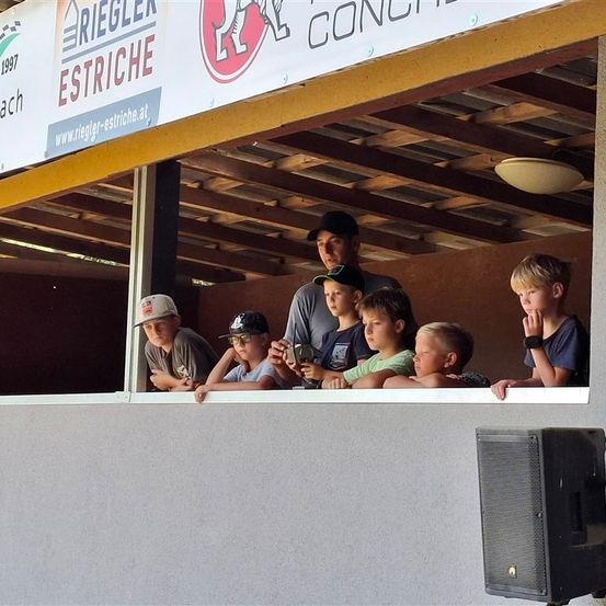 Mehrere Kinder und Erwachsene schauen von einer Kabine aus, auf der ein Banner steht, das 'RIEGLER ESTRICHE' besagt. Ein Lautsprecher befindet sich an der Wand rechts.