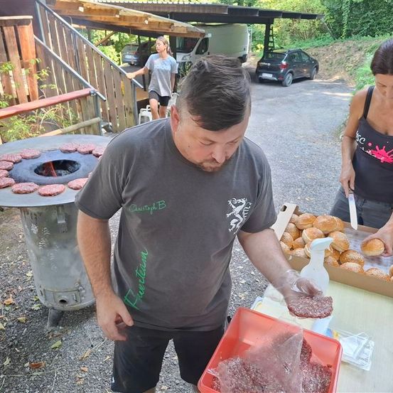 Ein Mann bereitet Burger vor einem Grill zu, mit Brötchen und Patties in einer Box bereit. Eine Frau schneidet im Hintergrund Burger.