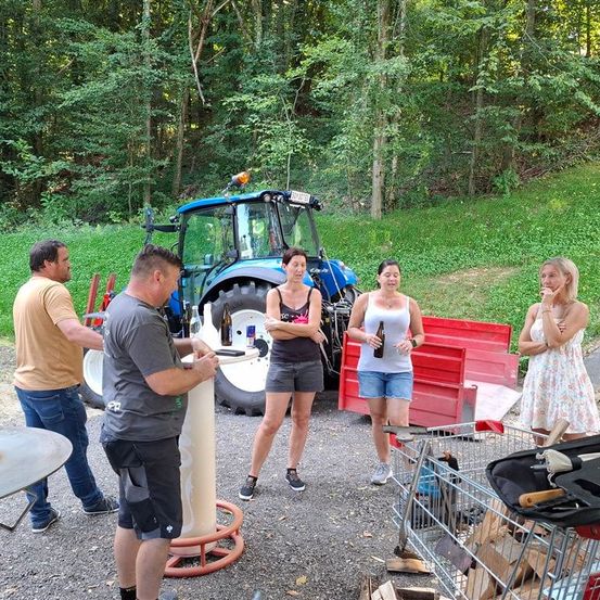 Eine Gruppe von Menschen steht draußen in der Nähe eines Traktors, wobei eine Person eine Flasche hält. In der Nähe befinden sich Werkzeuge und ein Tisch.