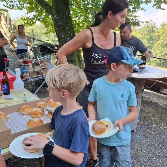 Eine Familie genießt ein Outdoor-Barbecue, zwei Jungen halten Hamburger, eine Frau vor ihnen und ein Mann am Grill im Hintergrund.