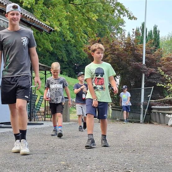 Eine Gruppe junger Jungen und ein Mann gehen auf einem Kiesweg, möglicherweise auf dem Weg zu einer Veranstaltung im Freien. Sie tragen Freizeitkleidung und Sneakers. Hinter ihnen befindet sich ein Zaun, Bäume und ein Haus.