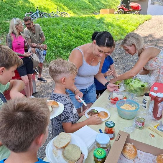 Ein Familienpicknick mit mehreren Personen um einen Tisch, eine Frau bereitet einen Salat zu und Kinder genießen ihr Essen.
