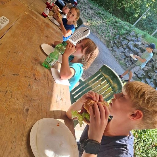 Eine Gruppe von Kindern isst im Freien an einem Picknicktisch, ein Junge isst einen Hamburger, ein anderer isst ein Sandwich und ein Mädchen trinkt aus einer Flasche.