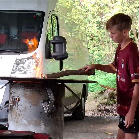 Ein Junge steht neben einem Tisch mit einem brennenden Stock, in der Nähe eines weißen Lieferwagens mit einem Feuer im Inneren und einem Spiegel an der Seite.