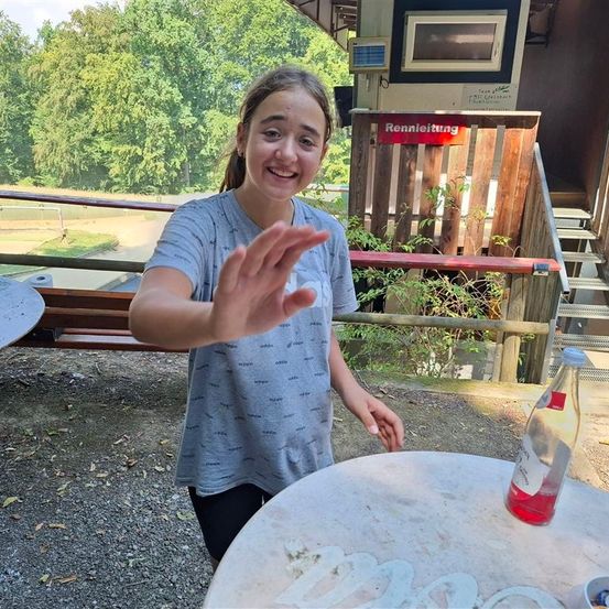 Ein junges Mädchen steht lächelnd in der Nähe eines Tisches mit einer Flasche roter Flüssigkeit, mit einem Holzzaun und Treppen hinter ihr.
