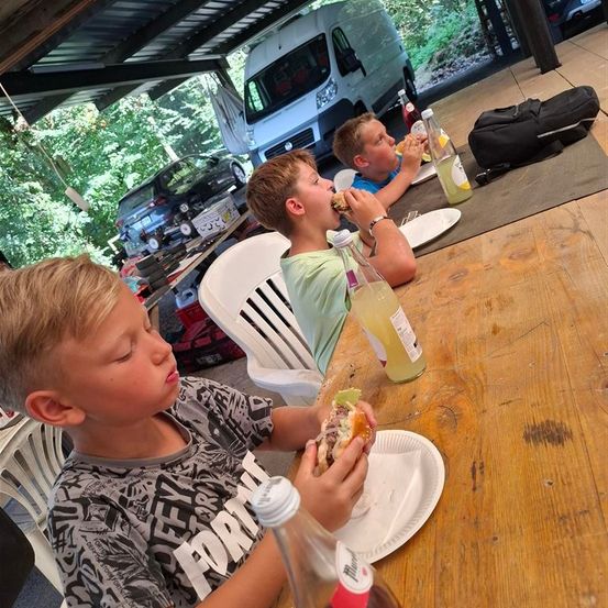 Zwei Jungen sitzen an einem Tisch und essen Sandwiches. Einer hat einen Burger und der andere ein Sandwich mit einem grünen Gemüse. Flaschen und Teller sind auf dem Tisch. Hinter ihnen steht ein Van und eine schwarze Tasche.