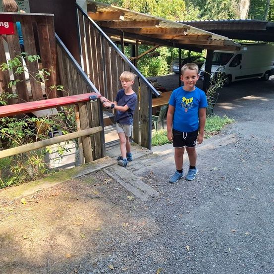 Zwei Jungen stehen vor einer hölzernen Struktur mit Treppen und einem Handlauf. Hinter ihnen stehen ein Tisch und Stühle unter einem Vordach. Ein Lieferwagen ist auf einem nahen Parkplatz abgestellt.