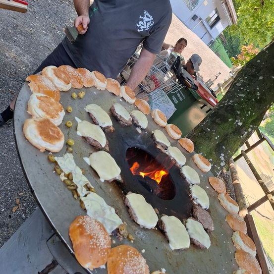 Ein Mann kocht zahlreiche Burger auf einem großen Grill mit Feuer darunter. Er steht neben einem Baum und Leute sitzen um einen Tisch herum.